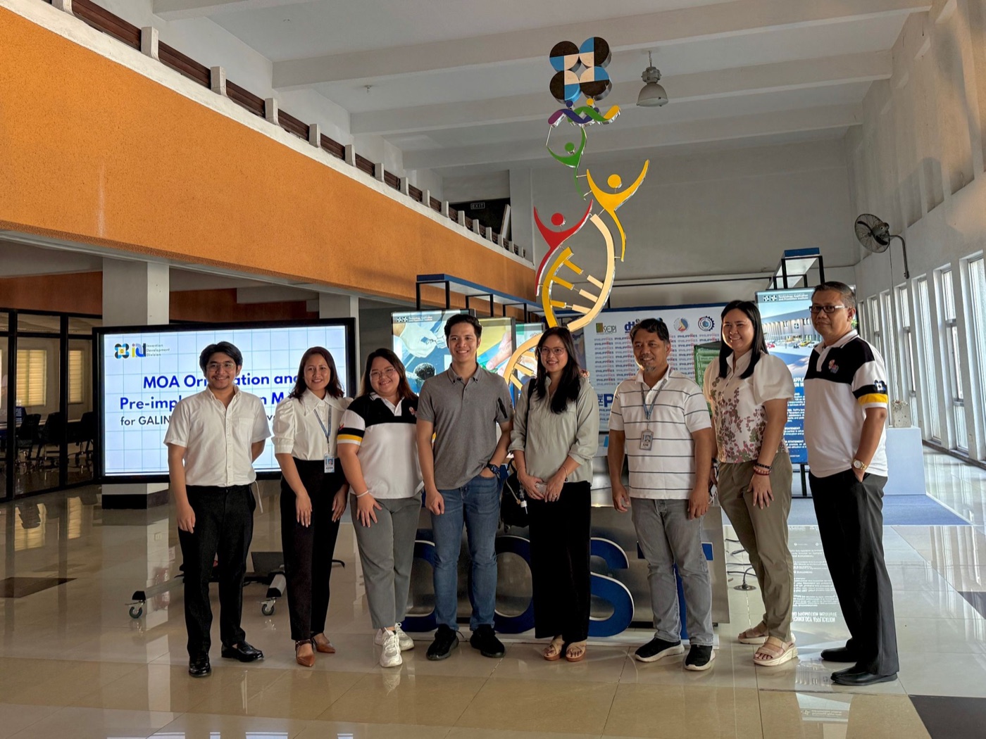Nine people standing together in the DOST lobby beneath a coloured DNA-helix sculpture, in front of a screen reading “MOA Orientation and Pre-implementation Meeting”.